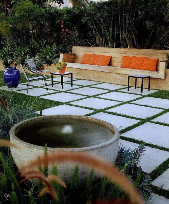 Moderne tuin met betonnen terrastegels, gras en een houten bank met oranje kussens.