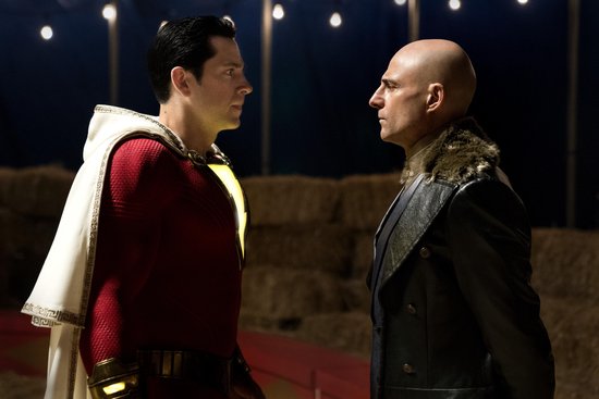 Shazam, in rood pak en witte cape, kijkt naar een kale man in een donkere jas met bontkraag.