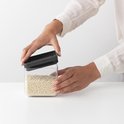 Des mains ouvrent une boîte de rangement carrée et transparente avec un couvercle gris foncé, remplie de couscous perlé.