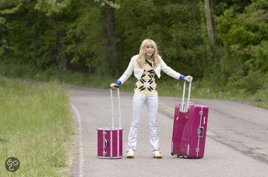 Miley Cyrus staat op een weg met twee roze koffers.