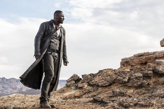 Idris Elba in een lange zwarte leren jas en westerse kleding staat op een rotsachtige heuvel met een dramatische hemel.