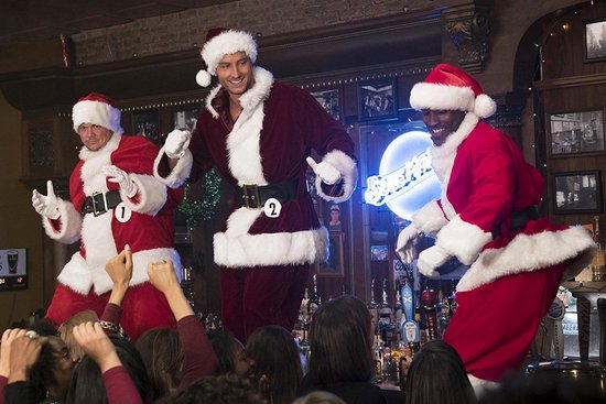Drie mannen in kerstmanpakken, twee met nummers op hun kostuum, staan op een podium.