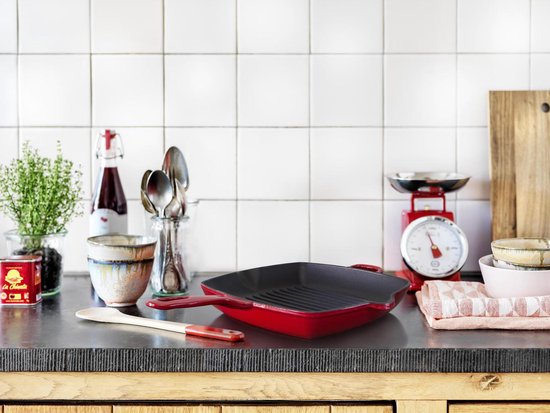 Rode gietijzeren grillpan op een keuken aanrecht, naast kookgerei en weegschaal.