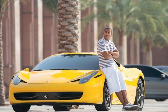 Een man in een gestreept shirt en witte shorts leunt tegen een gele Ferrari.
