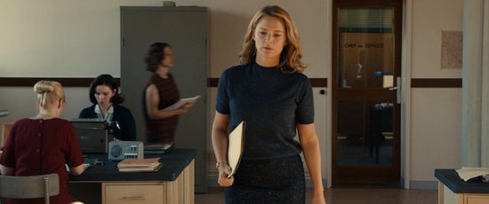 Een vrouw met blond haar in een donkerblauw gestreept shirt en rok, loopt met een map in een kantoor.
