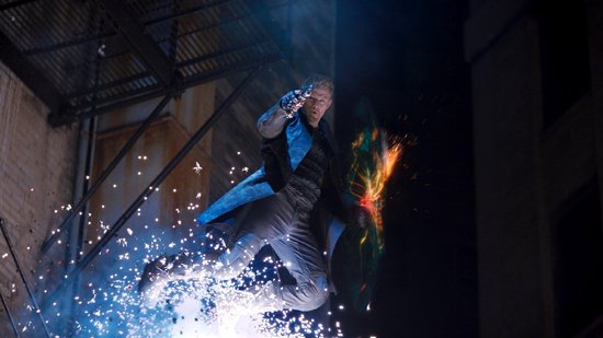 Een man in een blauwe jas springt in de lucht, met vuurwerk en lichteffecten om hem heen.