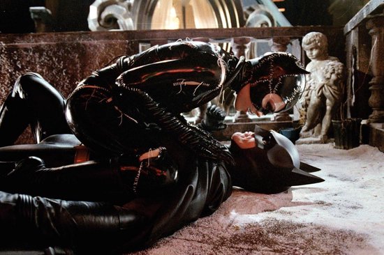 Catwoman, in een glanzend zwart pak, zit bovenop Batman, die op de grond ligt. Ze bevinden zich in een besneeuwde, donkere omgeving.