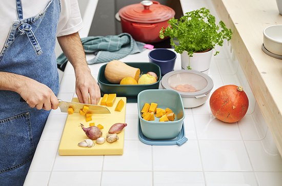 Un homme coupe des cubes de citrouille sur une planche à découper jaune. Des récipients vert clair, bleu clair et vert, une citrouille et une pomme sont sur le comptoir.