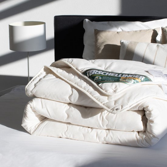 Opgevouwen off-white 4-seizoenen dekbed met groen label, op een bed met kussens en lamp.