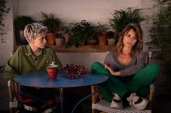 Twee vrouwen zitten aan een ronde tafel met planten. Eén met kort blond haar in groen, de ander met bruin haar in groen trainingspak.