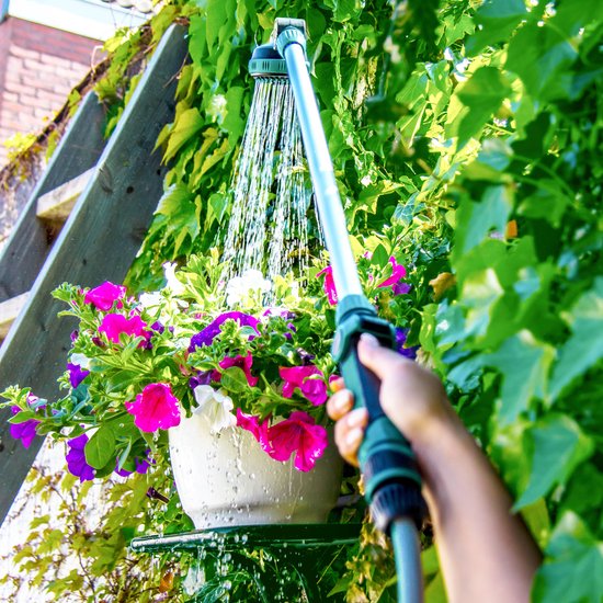 Een hand houdt een lange zilveren sproeilans met groene accenten vast, gericht op een pot met kleurrijke bloemen, tegen een groene achtergrond.