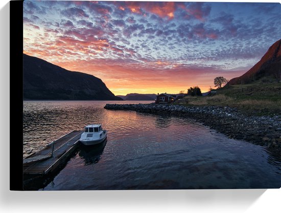 Een wit vissersbootje ligt aangemeerd bij een houten pier, met huizen op de oever en een dramatische zonsondergang met gekleurde wolken.