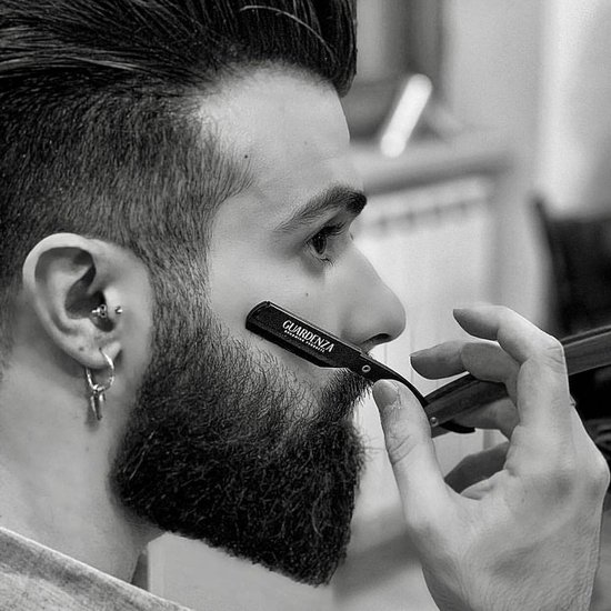 Zwart-wit foto van een man die zich scheert met een scheermes met houten handvat. Hij heeft een baard en oor piercings.