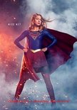 Supergirl in blauw pak met rood masker en cape, staat poseerend met handen in de zij.
