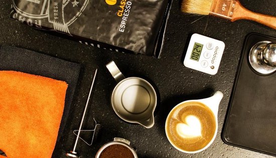 Koffieaccessoires: een kopje cappuccino met latte art, een portafilter, een maatbeker, een thermometer, een borstel en een weger.