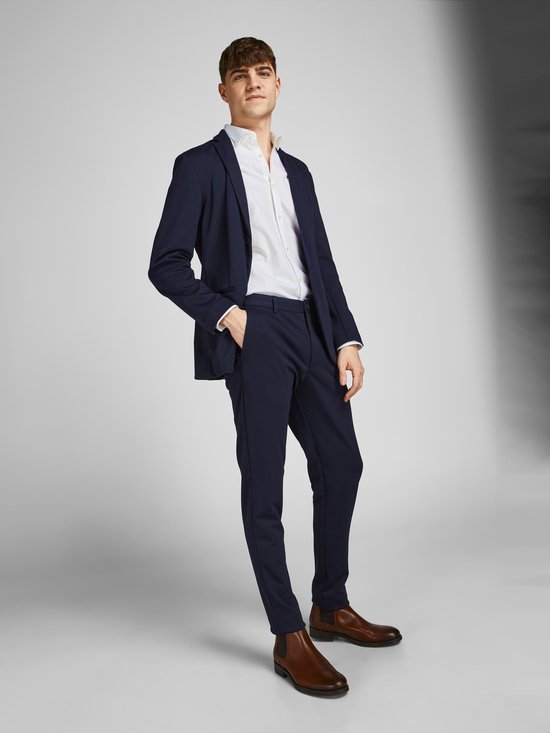 Man met donkerblauwe blazer en broek, wit overhemd en bruine schoenen.