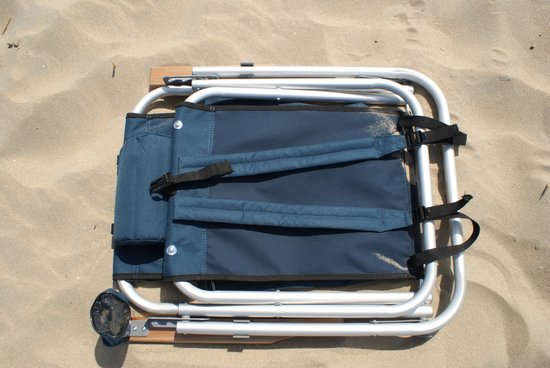Ingeklapte donkerblauwe campingstoel met zilverkleurig frame, liggend op zand. Met rugzakbanden.