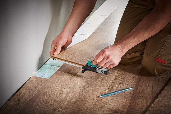Een persoon met beige broek knielt op een houten vloer en gebruikt een profielmal om de omtrek te meten.