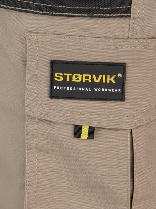 Salopette beige avec une étiquette STØRVIK "PROFESSIONAL WORKWEAR" et une boucle jaune et noire sur la poche.
