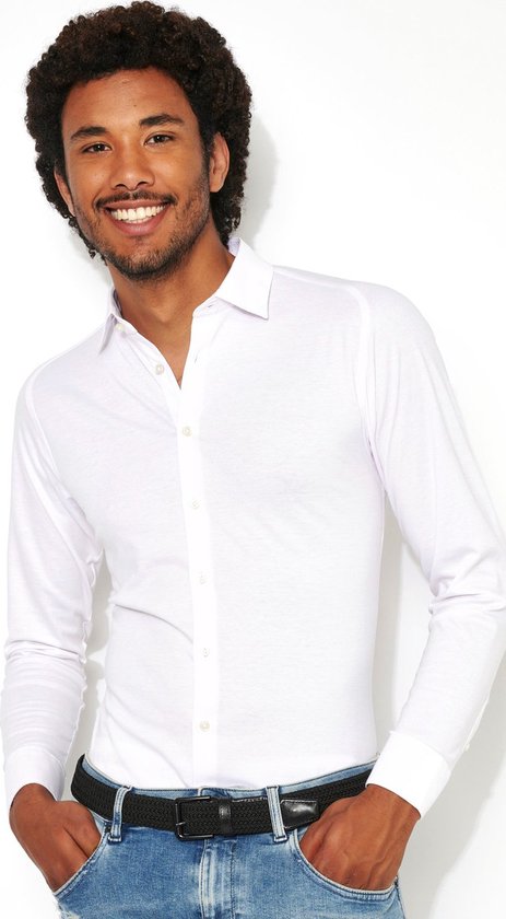 Man met krullend haar, lachend, draagt een wit overhemd met lange mouwen en een zwarte riem met jeans.