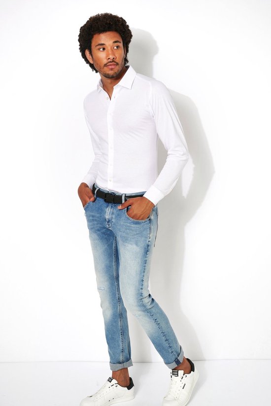 Man met krullend haar, draagt een wit overhemd met lange mouwen, een zwarte riem en een lichte jeans, sneakers.