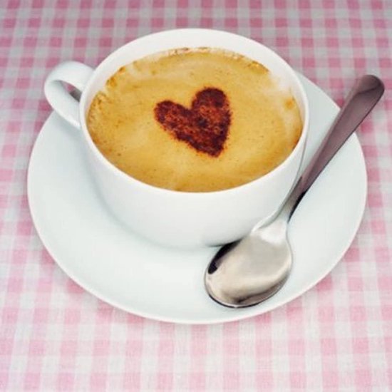 Koffiekopje met hart-patroon op melkschuim en een lepel op een roze-wit geblokt tafelkleed.