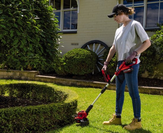 Een vrouw in jeans en grijze T-shirt maait een gazon met de rode Einhell accugrasmaaier.