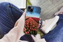 Handen houden een iPhone 13 hoesje vast met een veld met roze en rode tulpen. De persoon draagt een roze trui en spijkerbroek.