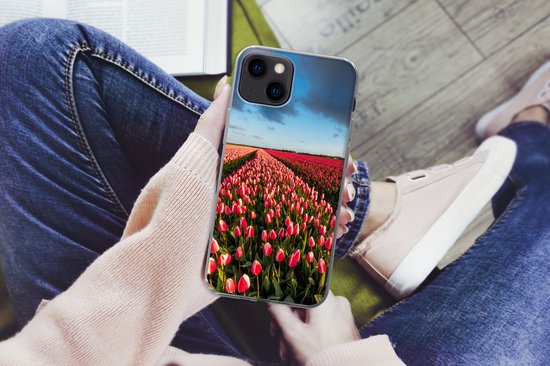 Handen houden een iPhone 13 hoesje vast met een veld met roze en rode tulpen. De persoon draagt een roze trui en spijkerbroek.