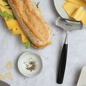 Een Boska kaasschaaf met zwart handvat en een stokbrood met kaas en rucola op een bord.