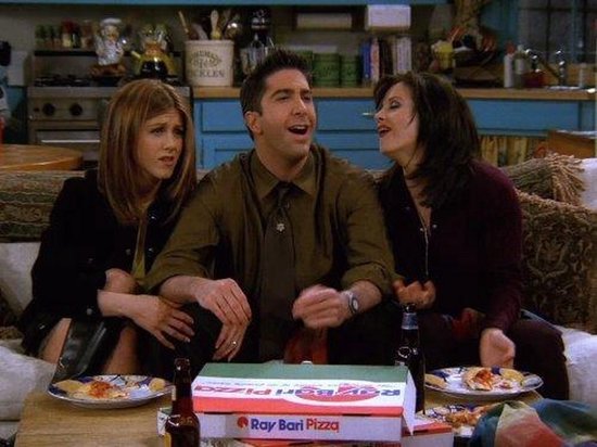 Drie vrienden zitten op een bank en kijken naar een doos pizza met daarop 'Ray Bari Pizza'.