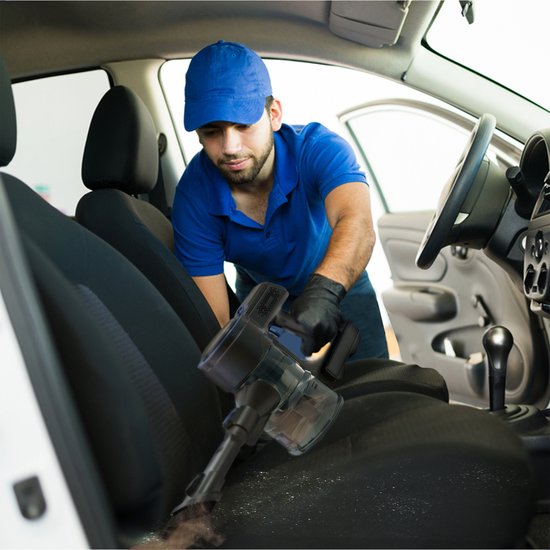 Man met blauwe pet en polo stofzuigt autostoel met zwarte draadloze stofzuiger.
