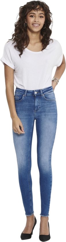 Vrouw met golvend bruin haar, draagt blauwe skinny jeans met rafelige zoom en witte opgerolde top, zwarte pumps.
