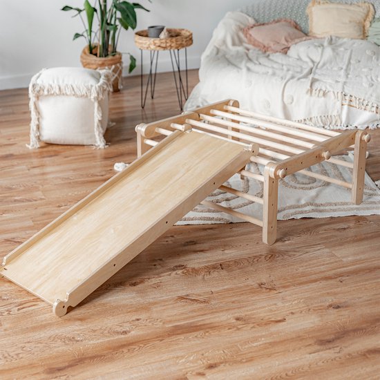 Een houten klimdriehoek met een glijbaan staat op een houten vloer, in een woonkamer met planten en een bed.