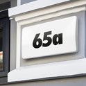 Huisnummer '65a' op een verlichte witte plaat, gemonteerd op een bakstenen muur.