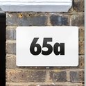 Verlichte witte huisnummerplaat '65a' gemonteerd op een bakstenen muur.