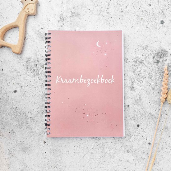 Roze kraambezoekboek met de tekst 'Kraambezoekboek' in sierlijke letters. Sterren en een maan sieren de omslag. Naast het boek liggen een houten giraf en een gedroogd grasplukje.