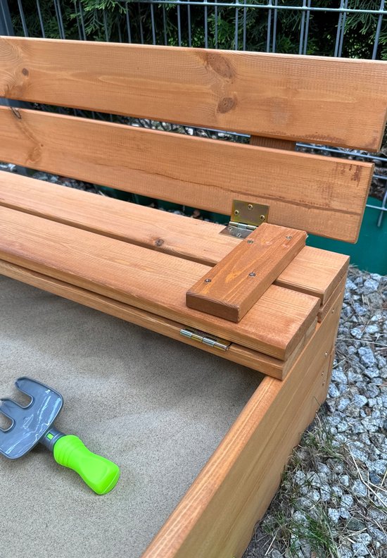 Detailopname van de scharnieren op de houten bank van een zandbak. Een graafspeeltje ligt in het zand.