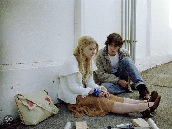 Een jonge vrouw met blond haar en een man zitten op de grond. Een tas met rode harten staat naast de vrouw.
