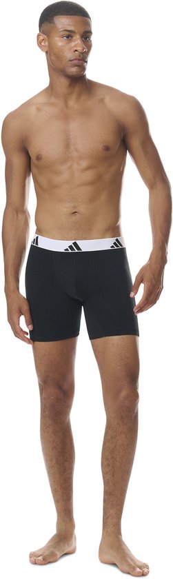 Model draagt zwarte boxershort met witte adidas tailleband, staand.