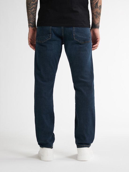 Man met tatoeages op armen, staand met de rug naar de camera in een zwarte t-shirt en donkerblauwe jeans.