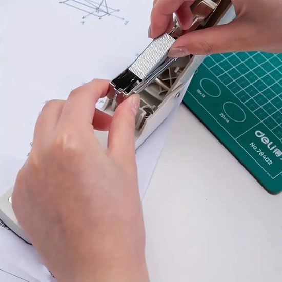 Handen vullen een nietmachine met zilverkleurige nietjes, met papieren en een groene snijmat op de achtergrond.