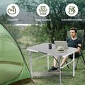 Man zit aan een zilverkleurige campingtafel met laptop en hoed, naast een tent.