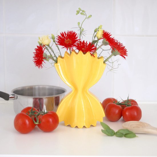 Gele, strikvormige vaas gevuld met rode en witte bloemen, op een aanrecht met tomaten.