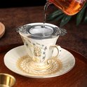 Zilverkleurige theezeef met decoratieve vleugel handvatten, in een antiek wit theekopje met gouden decoratie.