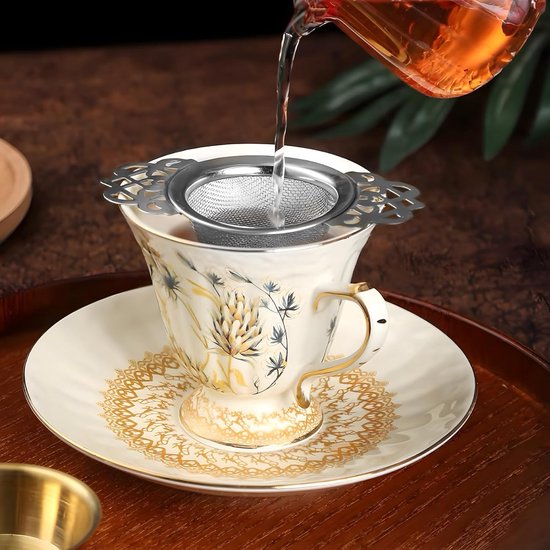 Zilverkleurige theezeef met decoratieve vleugel handvatten, in een antiek wit theekopje met gouden decoratie.