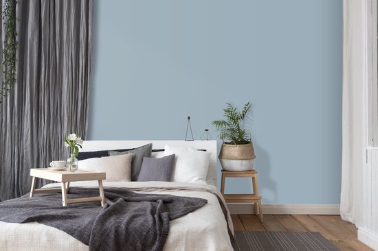Een slaapkamer met een poederblauwe muur, een wit bed met grijs beddengoed en decoratie, een plant op een krukje.