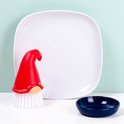 Rode gnome-afwasborstel met witte haren, naast een wit bord en een blauw bakje.