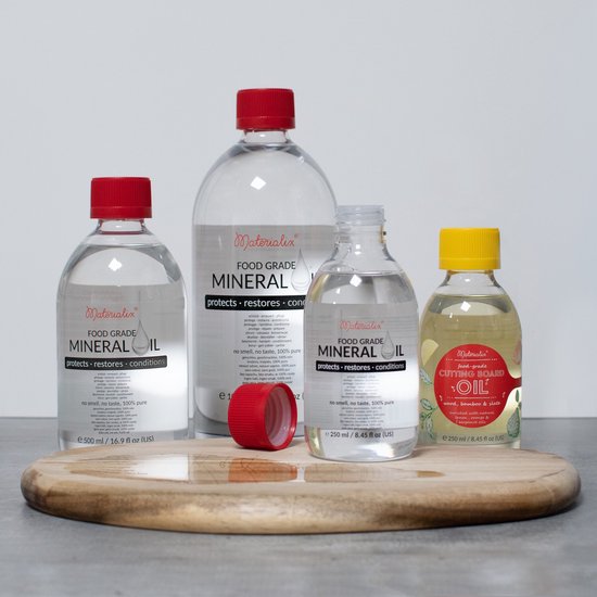 Vier flessen Materialix Food Grade Mineral Oil en een fles snijplankolie op een houten snijplank.