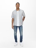 Man met korte mouwen in lichtgrijs, open overhemd over een wit T-shirt en distressed jeans.